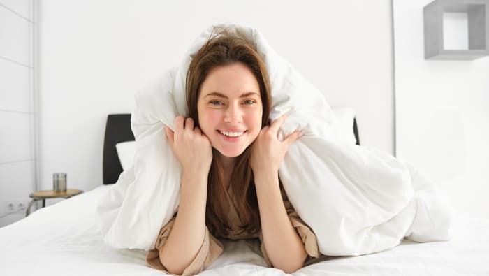 Mulher sorrindo sobre cama com roupa de cama branca, representando conforto, bem-estar, qualidade do sono e experiência ligada ao mercado de colchões
