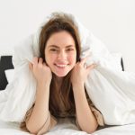 Mulher sorrindo sobre cama com roupa de cama branca, representando conforto, bem-estar, qualidade do sono e experiência ligada ao mercado de colchões